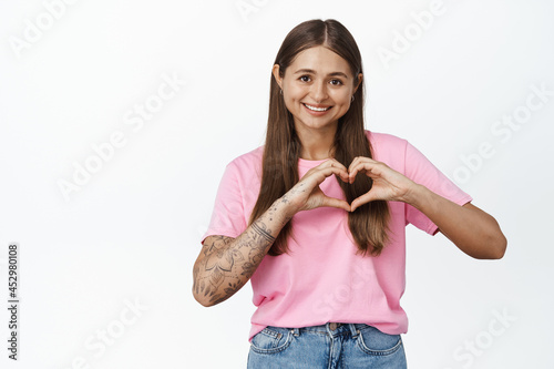 Romantic girl shows heart sign and smiles at camera, stands in pink tshirt with jeans, white background