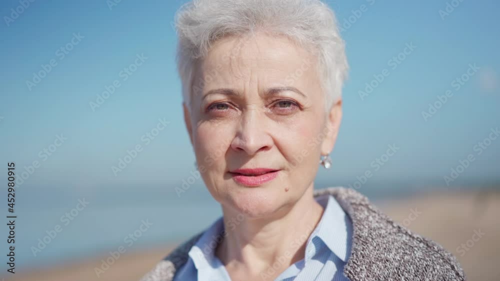 Head and shoulders closeup portrait of senior woman with white short hair looking at camera, smiling and laughing on beach. Mature female posing on sea coast. Travel and happy retirement concepts Head and shoulders closeup portrait of senior woman with white short hair looking at camera, smiling and laughing on beach. Mature female posing on sea coast. Travel and happy retirement concepts