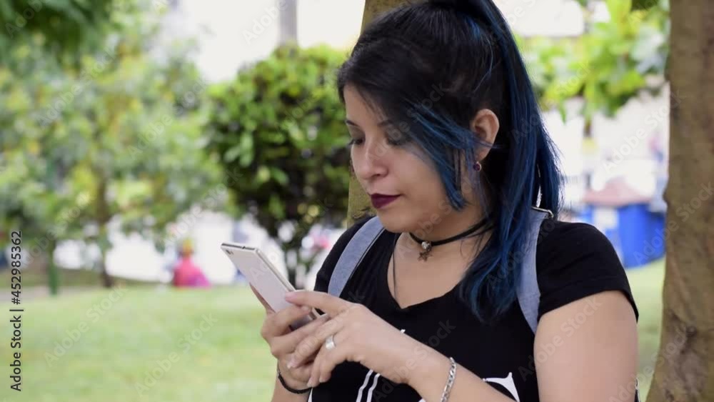 Beautiful young woman smiling. blue haired college girl looking at her social media in a park, next to a tree. girl looking at her cell phone in a park. Technology, communication and education concept Beautiful young woman smiling. blue haired college girl looking at her social media in a park, next to a tree. girl looking at her cell phone in a park. Technology, communication and education concept