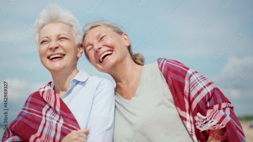 Zoom in slow motion shot of happy senior girlfriends wrapped in checkered blanket laughing joyfully while walking on sea beach. Two cheerful retired women having fun together, candid emotions Zoom in slow motion shot of happy senior girlfriends wrapped in checkered blanket laughing joyfully while walking on sea beach. Two cheerful retired women having fun together, candid emotions