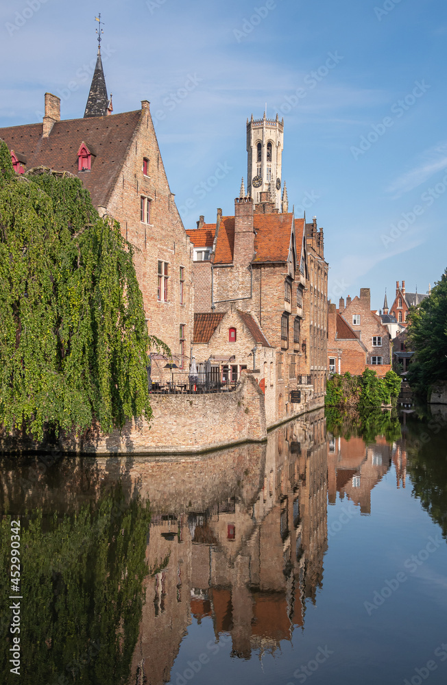 Brugge, Flanders, Belgium - August 4, 2021: Sunlit Belfry towers over brown brick back facades, mirrored in quiet Dijver canal, with green foliage in corners under blue sky. 