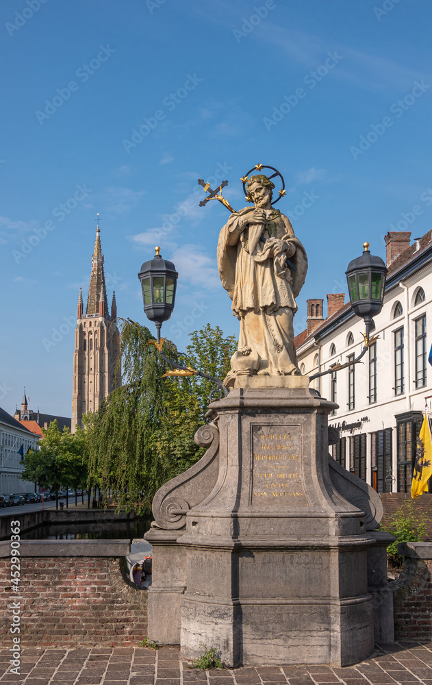 Fototapeta premium Brugge, Flanders, Belgium - August 4, 2021: Closeup of Nepomucenus stone statue on his bridge with Notre Dame cathedral tower in back under blue sky. Backdrop of green foliage and facades.