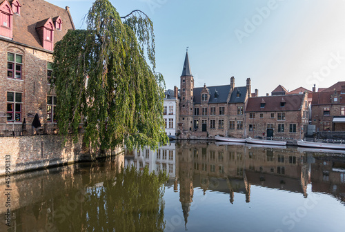 Brugge, Flanders, Belgium - August 4, 2021: Quiet Dijver canal reflects brown stone Huidevettershuis and boats under light blue sky and green foliage on side.