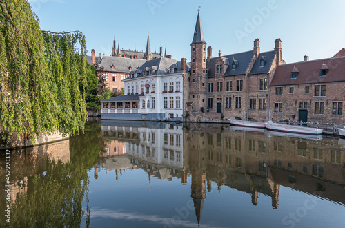 Brugge, Flanders, Belgium - August 4, 2021: Quiet Dijver canal reflects brown stone Huidevettershuis and white painted Duc de Bourgogne restaurant under light blue sky and green foliage on side. photo