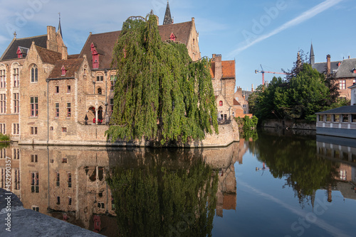 Brugge, Flanders, Belgium - August 4, 2021: Quiet Dijver reflects sunlit brown brick buildings of Wollestraat with green foliag in center and back. 