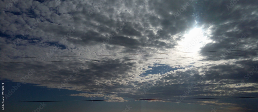 A panorama of a cloudy sky over the sea surface. Smooth lime-colored water and uniform clouds create a calm, serene mood of the landscape. A white disk of the sun peeps through fluffy silvery clouds. A panorama of a cloudy sky over the sea surface. Smooth lime-colored water and uniform clouds create a calm, serene mood of the landscape. A white disk of the sun peeps through fluffy silvery clouds.