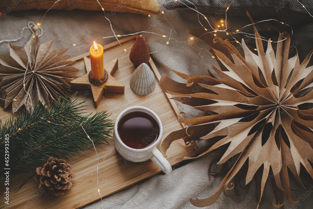 Warm cup of tea with christmas lights, stars, pine trees, candle and pillows on soft bed. Flat lay. Cozy home. Winter hygge. Festive holiday decorations in scandinavian room. Atmospheric moment Warm cup of tea with christmas lights, stars, pine trees, candle and pillows on soft bed. Flat lay. Cozy home. Winter hygge. Festive holiday decorations in scandinavian room. Atmospheric moment
