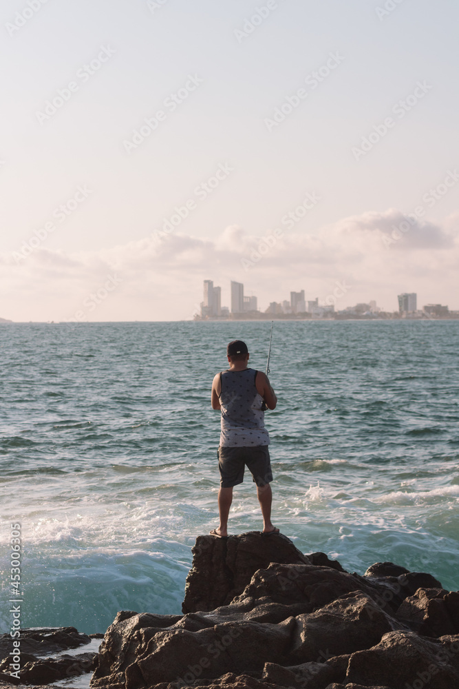 Man standing on a rock fishing in the ocean. City skyline in the background