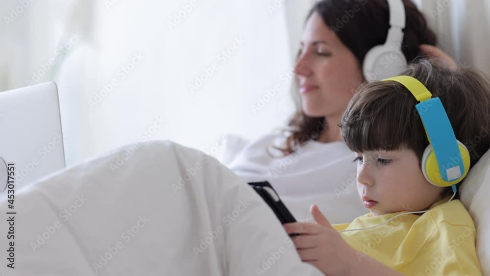 Lazy family spend day in bedroom wearing pajamas while working and learning online: mom and kid boy in headphones listen music looking at laptop and digital tablet screen lying in bed under blanket Lazy family spend day in bedroom wearing pajamas while working and learning online: mom and kid boy in headphones listen music looking at laptop and digital tablet screen lying in bed under blanket