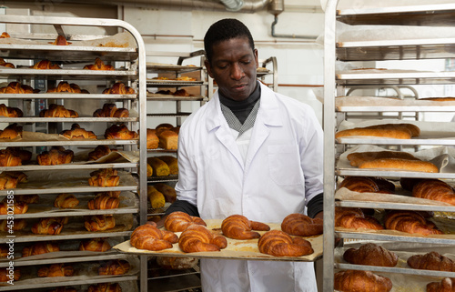 Portrait of african american baker working in bakehouse, putting tray with fresh bakery goods on rack trolley