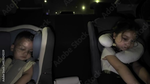Cute little siblings sleep in the back seat in a car seat at night. The car is on the road. The child is tired after a long journey by car. Uncomfortable sleeping position. Auto-pillow for sleeping