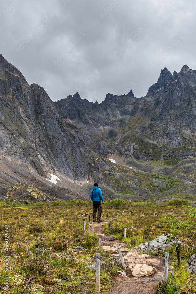 Hiker in backcountry campground of Grizzly Lake, Yukon. Amazing towering mountain peaks behind man wearing blue jacket, black, brown pants with camera. 