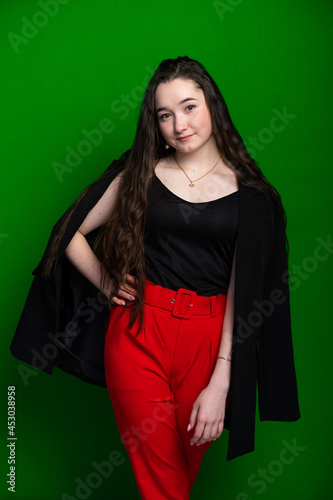 portrait of a girl. model in red trousers, black shoes and a jacket. long curly hair. colored green background.