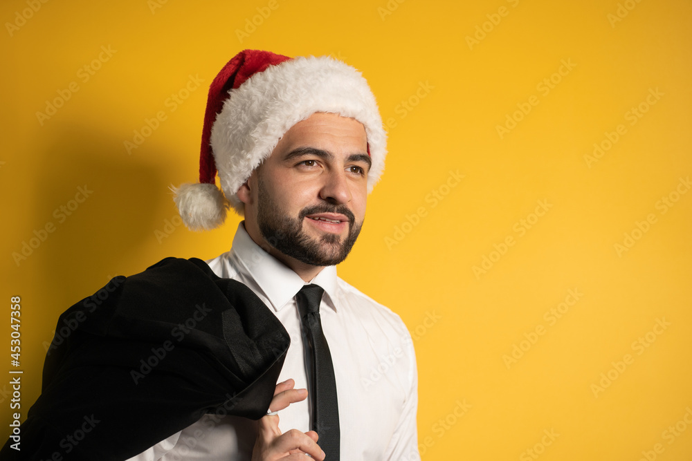 Portrait of a handsome Santa businessman in red santa hat posing holding his black jacket on his shoulder, hanging it behind looking cheerful at camera isolated on yellow background. Portrait of a handsome Santa businessman in red santa hat posing holding his black jacket on his shoulder, hanging it behind looking cheerful at camera isolated on yellow background.