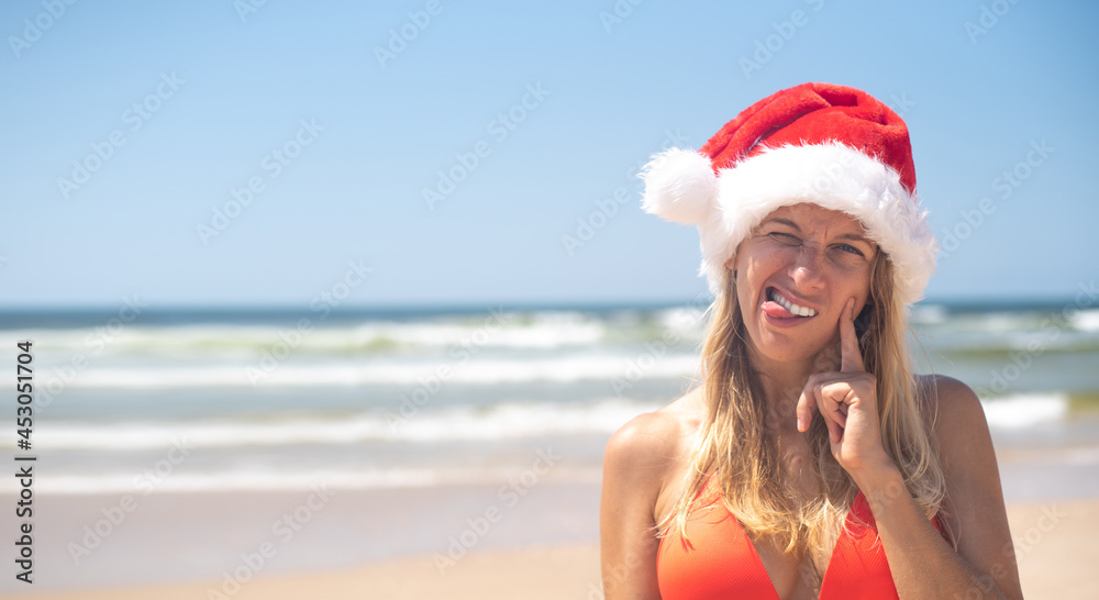 Obraz premium Girl wear red Santa hat stay on seashore points right with brooded expression and drained face. Travel on new year on sea in Christmas. Holiday and vacation on beach.