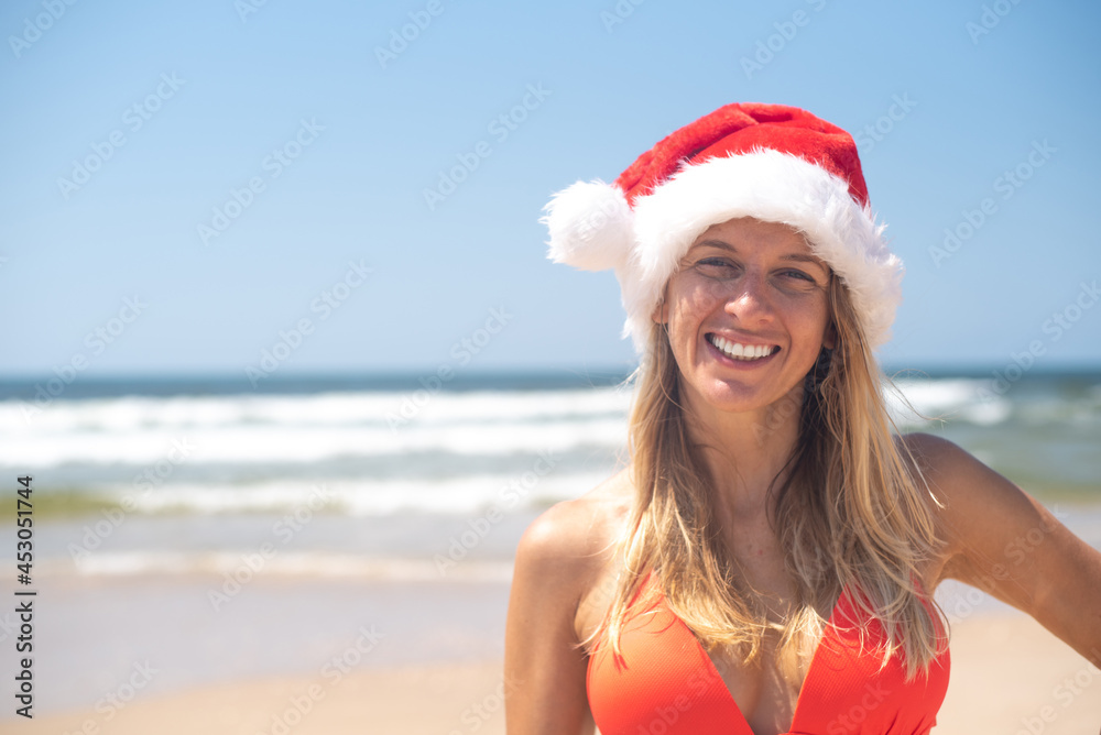 Obraz premium girl wear red Santa hat and red bikini with happy face stay on seashore background, space for text. Travel in new year and Christmas to sea, holiday and vacation on ocean.