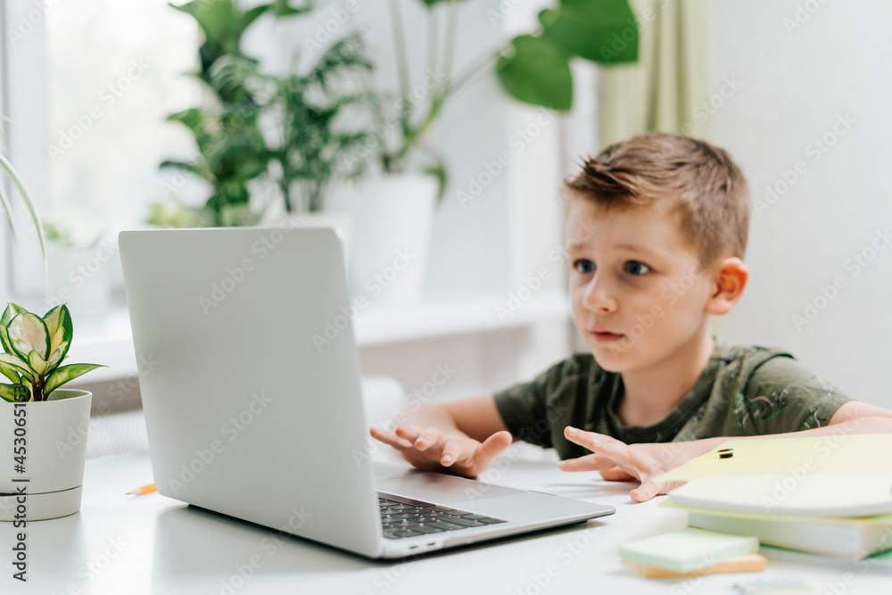 Distance learning online education. Caucasian smile kid boy studying at home with laptop and doing school homework. Surprised child siting with notebook, pencils and training books. Back to school.