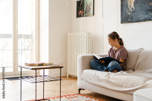 White preteen girl smiling and reading book while sitting on couch