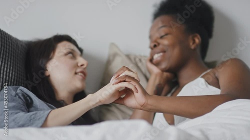 Medium shot with slowmo of multi-ethnic loving lesbian couple lying in bed and intertwining fingers while smiling to each other and chatting