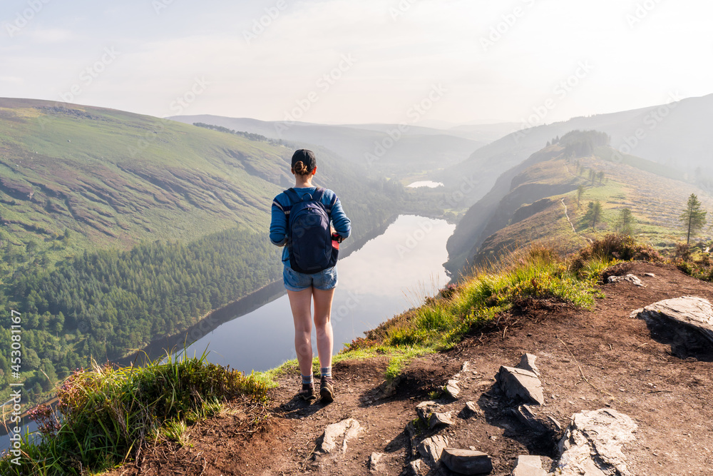 Female hiker enjoying the view from The Spinc trail over Glendalough Upper Lake on a hazy summer morning in Wicklow Mountains, Ireland.