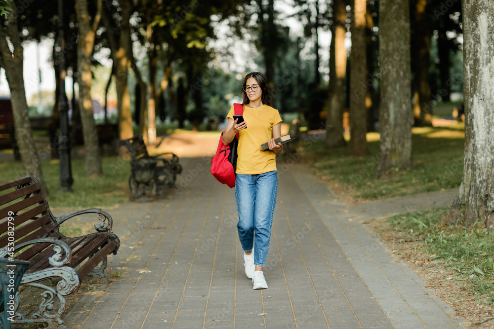 Fototapeta premium Portrait of a young student girl walking through the park with a backpack, a book and a phone in her hands