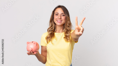 pretty thin woman smiling and looking happy, gesturing victory or peace and holding a piggybank photo