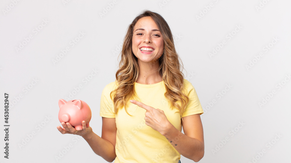 pretty thin woman smiling cheerfully, feeling happy and pointing to the side and holding a piggybank - obrazy, fototapety, plakaty 