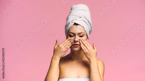 young woman with white towel on head applying exfoliating mask isolated on pink