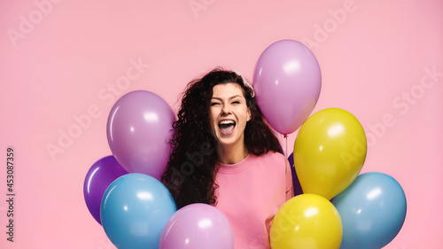 overjoyed woman with plenty of multicolored balloons isolated on pink