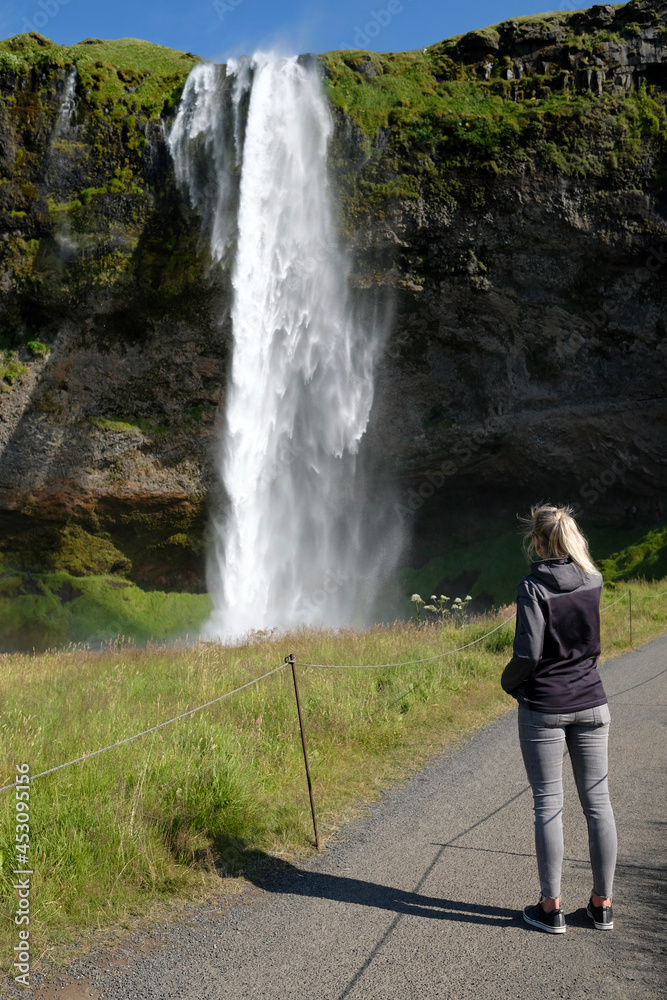 Woman seen from the back admiring Seljalandsfoss waterfall on the southern coast of Iceland on a sunny day