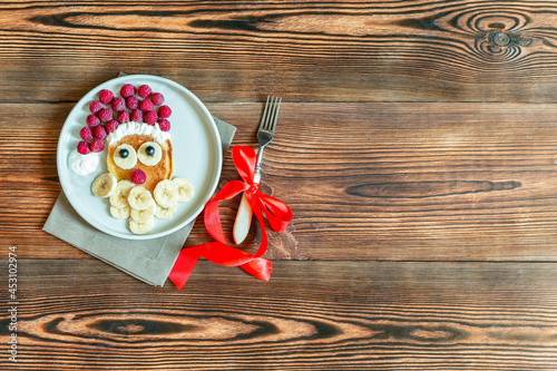Christmas Santa Claus shaped pancake with sweet fresh raspberry berry and banana on plate on wooden background for kids children breakfast. xmas food with new year decorations with copy space