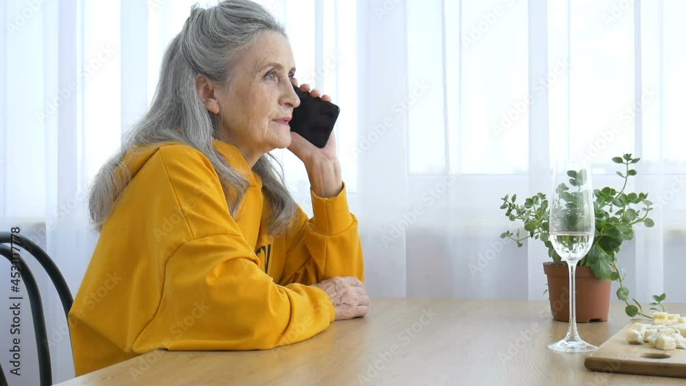 Beautiful old grandmother with grey hair and face with wrinkles is using smartphone, talking with someone and sitting at the table at home on window background, mother's day, happy retirement Beautiful old grandmother with grey hair and face with wrinkles is using smartphone, talking with someone and sitting at the table at home on window background, mother's day, happy retirement