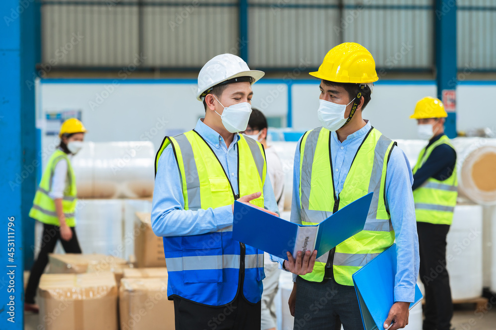 Team engineers and foreman wear a mask, hard hat, and vest. Standing, consult discuss industrial production management through data files with in mask factory. Employees working in the background. Team engineers and foreman wear a mask, hard hat, and vest. Standing, consult discuss industrial production management through data files with in mask factory. Employees working in the background.