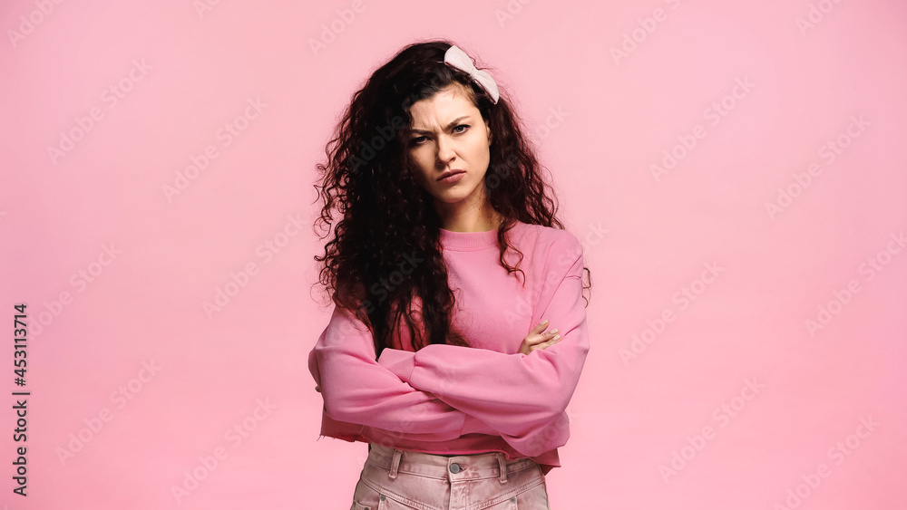 frowning woman looking at camera while standing with crossed arms isolated on pink frowning woman looking at camera while standing with crossed arms isolated on pink