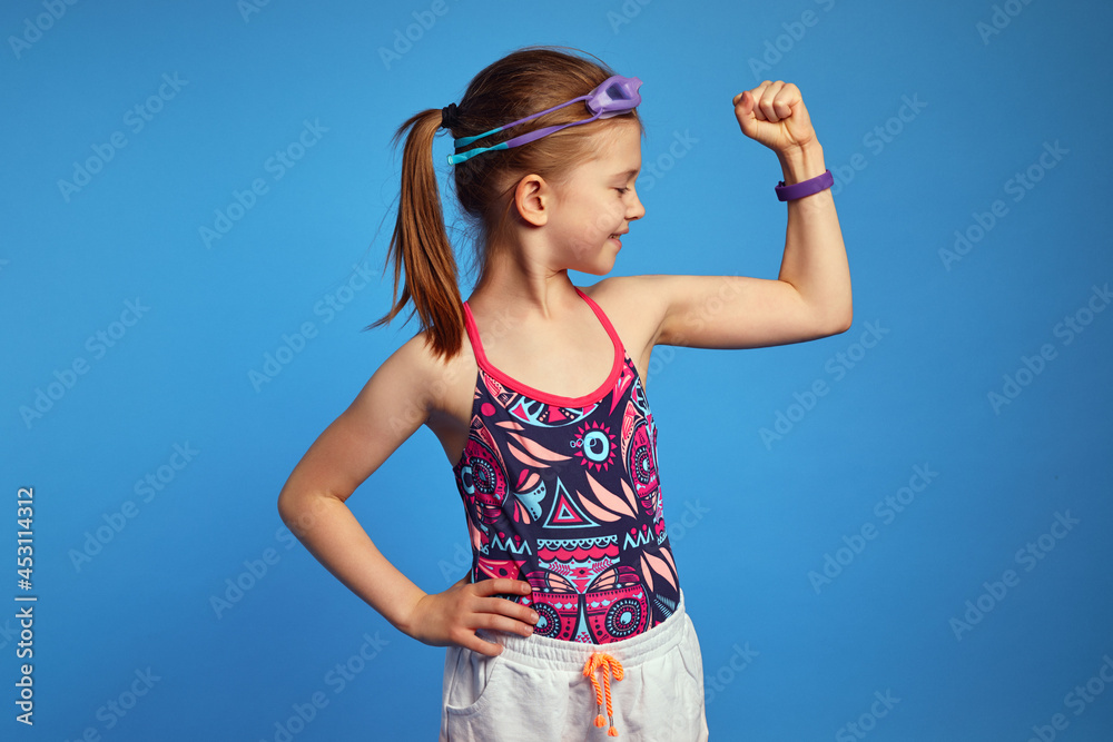 Little girl spends leisure time in water park, raises arm and looking at muscles, ready for swim, poses over blue background, wears goggles and swimsuit Little girl spends leisure time in water park, raises arm and looking at muscles, ready for swim, poses over blue background, wears goggles and swimsuit