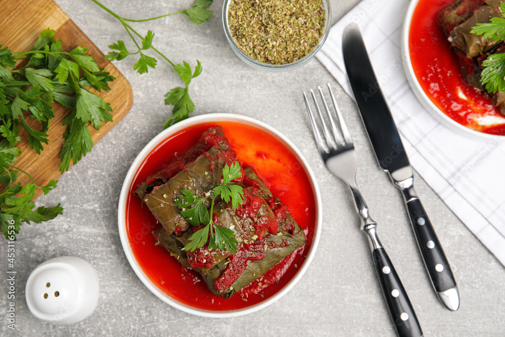 Plate of delicious stuffed grape leaves with tomato sauce, parsley and cutlery on light grey table, flat lay Plate of delicious stuffed grape leaves with tomato sauce, parsley and cutlery on light grey table, flat lay