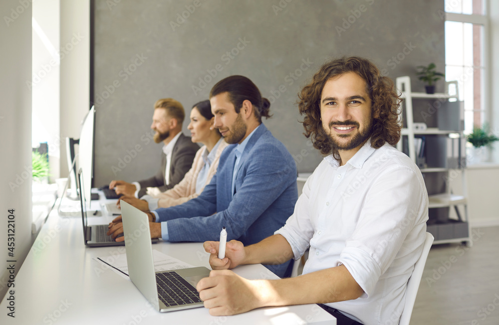 Group of business people working on modern desktop and laptop PCs. Portrait of happy handsome long haired man looking at camera while sitting at office desk with team of other employees and coworkers Group of business people working on modern desktop and laptop PCs. Portrait of happy handsome long haired man looking at camera while sitting at office desk with team of other employees and coworkers