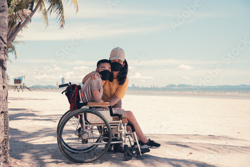 Mother and disabled teenager boy wearing face mask on outdoors public place like everyone else, The sea beach background, Lifestyle of handicapped vacation, Diverse people concept.