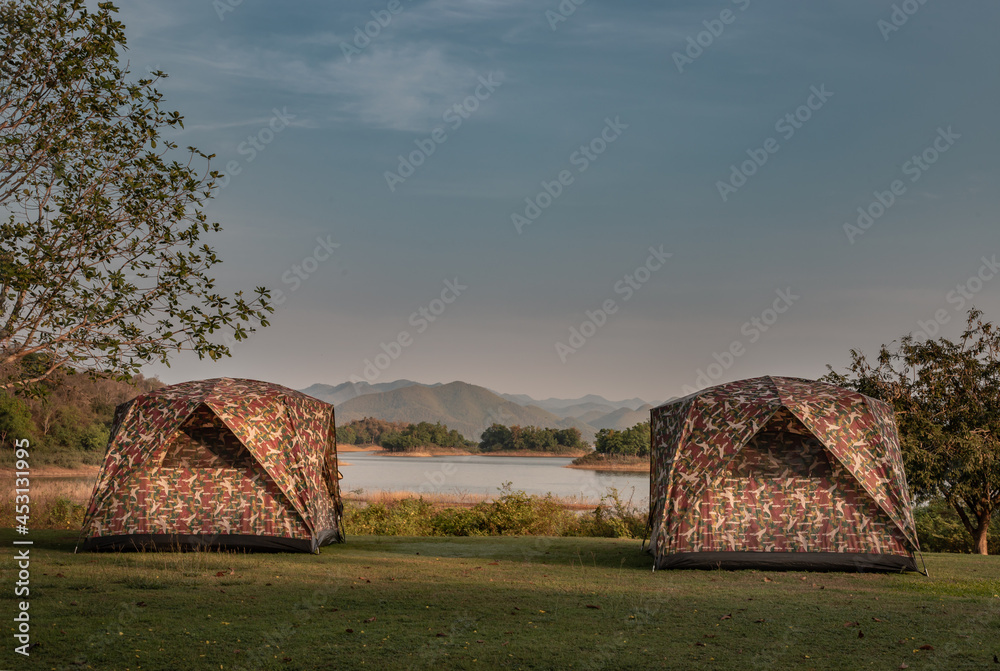 View of two camping tent with awning pitched on the dry ground Near beautiful big trees with branches at the campsite in lake shore with mountain range in background. Selective focus.