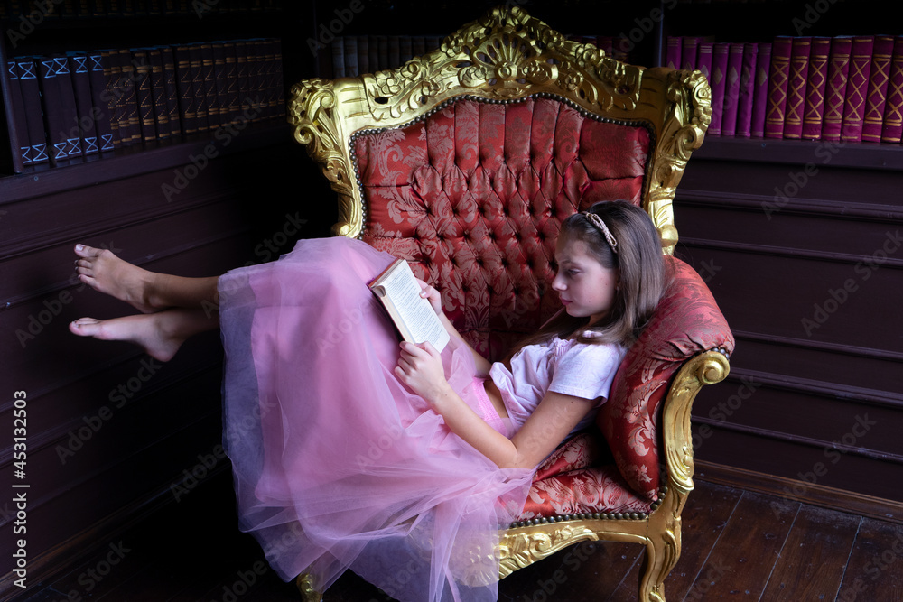 Lovely teen girl in pink tutuskirt reading book while sitting sideways on chic goldlined