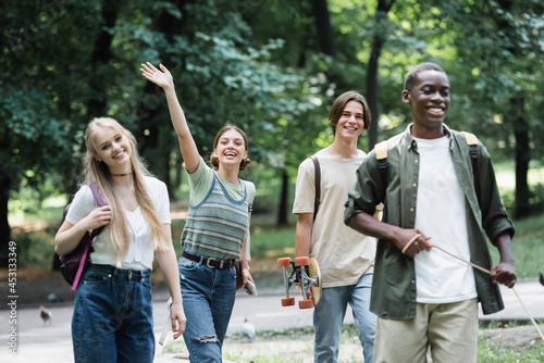 Smiling teenager waving at camera near interracial friends in park
