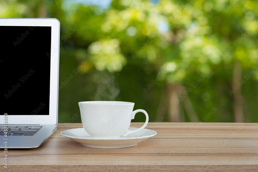 A white coffee mug and laptop is placed on old wooden top table with blurred green leaves, bokeh in garden background. Good morning or have a happy day and positive message concept. A white coffee mug and laptop is placed on old wooden top table with blurred green leaves, bokeh in garden background. Good morning or have a happy day and positive message concept.