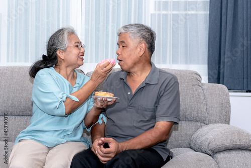 Asian elderly couple, are sitting on a sofa in home holding and eating donuts as a snack with happy together, to elderly and food concept.