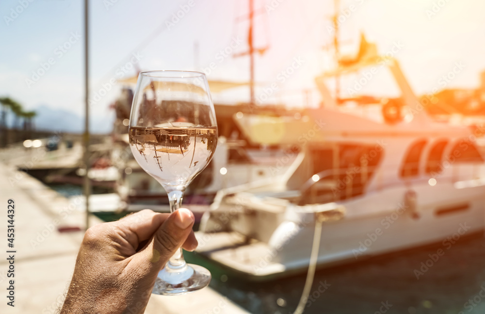 Obraz premium Glass of wine in hand. A glass of white wine against the backdrop of the Mediterranean sea and the port with yachts in a tourist town in the summer under sunlight. Summer, travel, lifestyle
