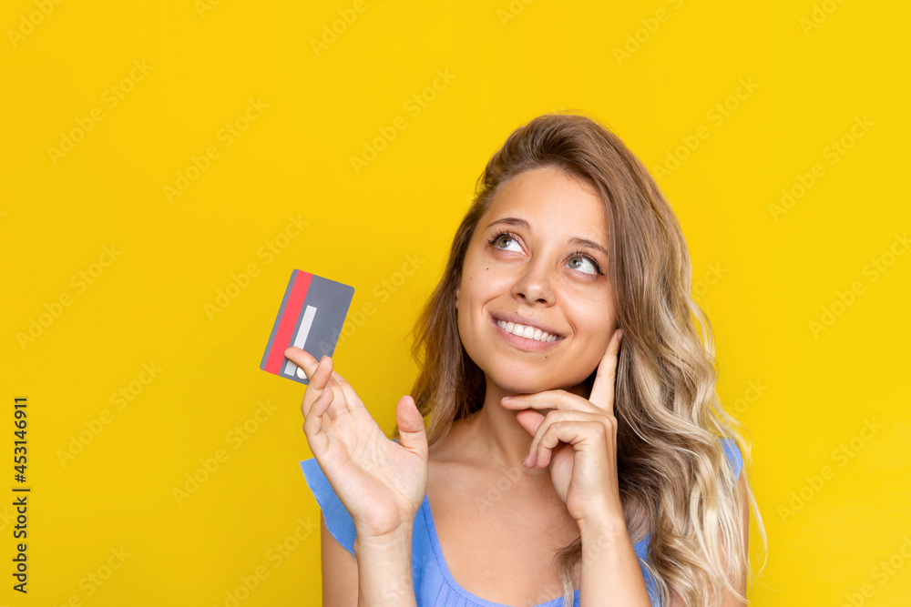 Close-up of a young blonde woman with wavy hair holding a plastic credit card in her hand thinking about where to spend the money looking up at empty copy space isolated on a color yellow background Close-up of a young blonde woman with wavy hair holding a plastic credit card in her hand thinking about where to spend the money looking up at empty copy space isolated on a color yellow background