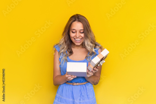 A young charmig amazed excited blonde woman with wavy hair in a blue dress is happy looking into an open gift box isolated on a color yellow background. A satisfied girl received a pleasant surprise