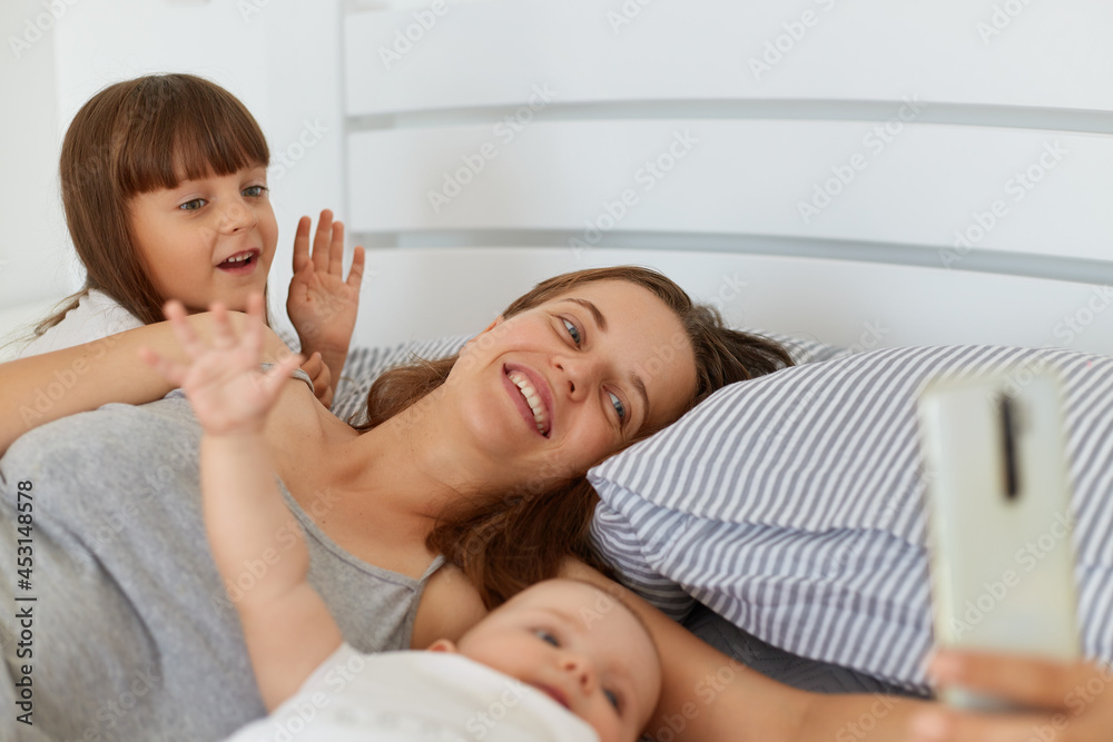 Happy positive female lying in bed with her children and holding smart phone in hands, people waving hands ans say hello, family blog, online conversation with followers.