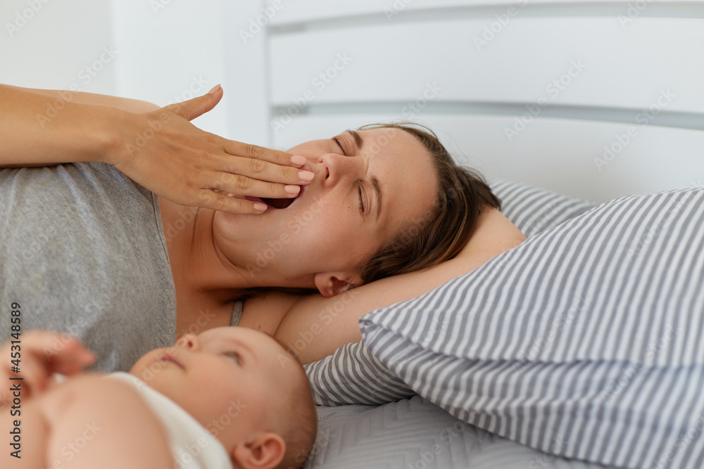 Indoor portrait of tired mother lying in bed with little baby girl or boy, yawning, covering mouth with palm, looks sleepy, having lack of energy and sleepless nights. Indoor portrait of tired mother lying in bed with little baby girl or boy, yawning, covering mouth with palm, looks sleepy, having lack of energy and sleepless nights.