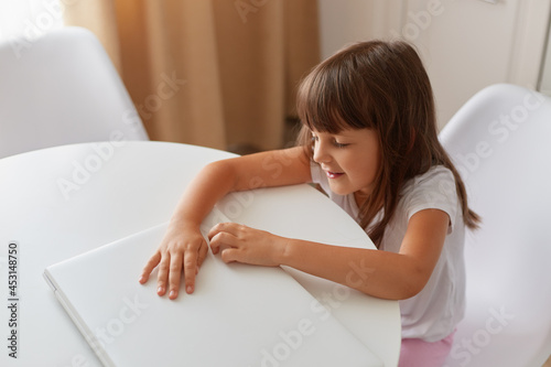Cute little girl is sitting at table with her white closed laptop, dark haired female child posing in light room wearing casual style closing, indoor shot.