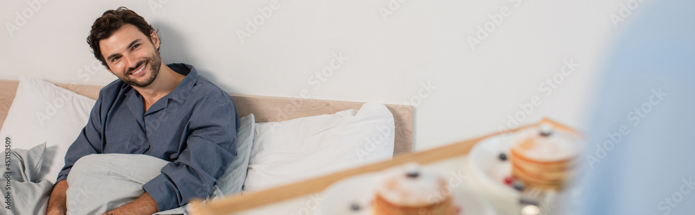 blurred man holding breakfast tray with pancakes near happy boyfriend in bed, banner blurred man holding breakfast tray with pancakes near happy boyfriend in bed, banner
